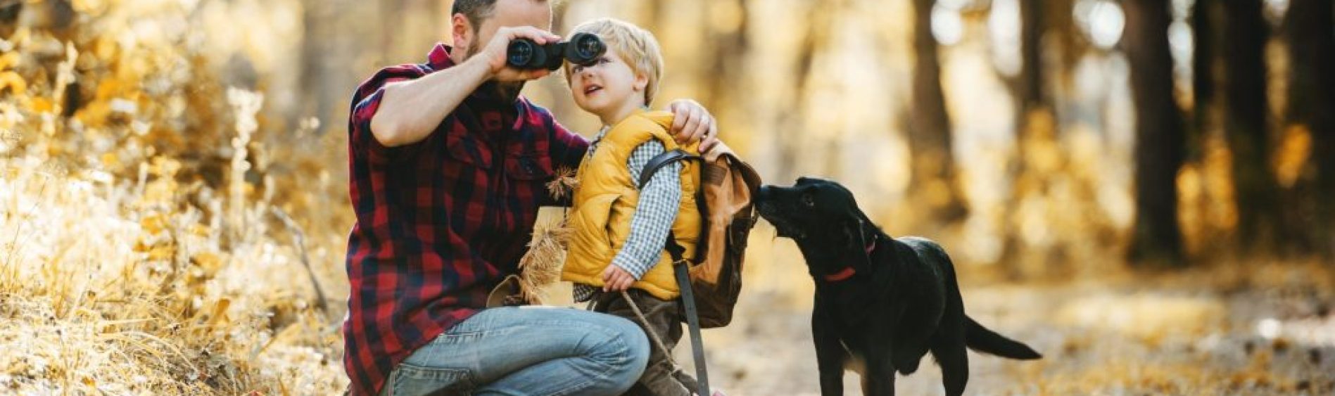 A mature father with a dog and a toddler son in an autumn forest, using binoculars.